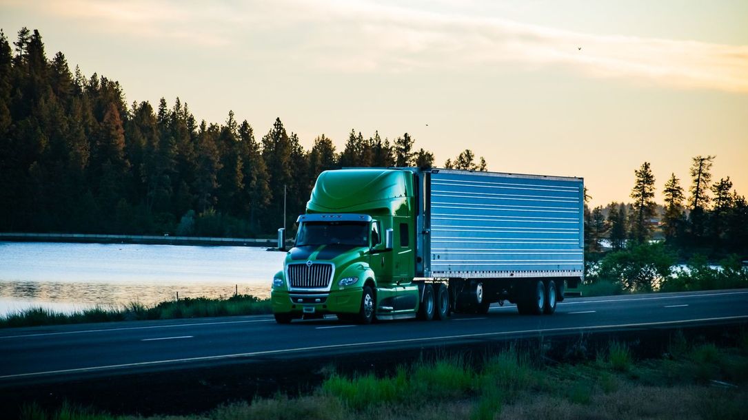 Green semi-truck driving on a road next to a lake with a forest in the background.