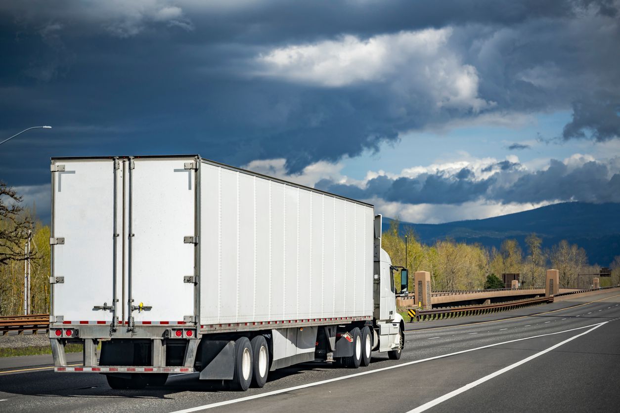 White semi-truck driving on a highway with mountains in the background under a cloudy sky.