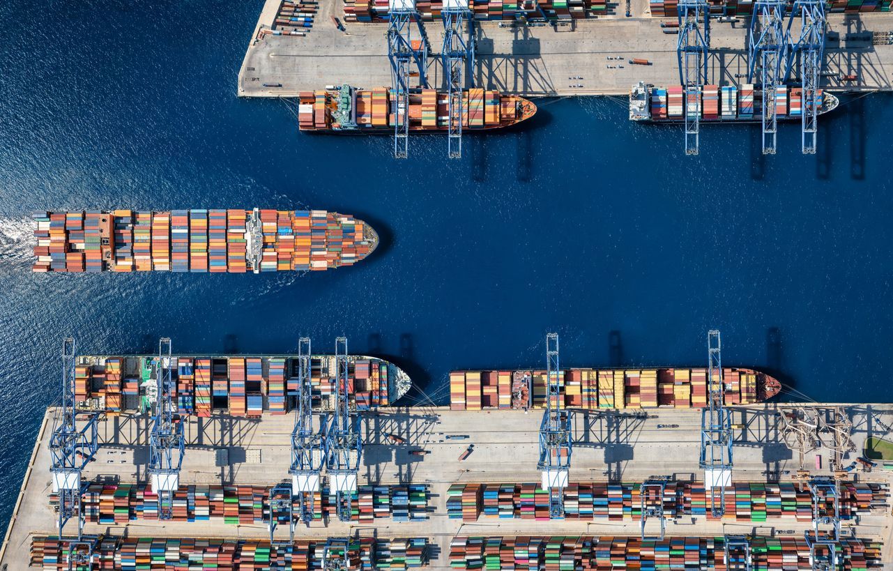 Aerial view of a busy harbor filled with cargo ships and cranes, with blue water and colorful containers.