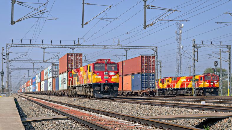 Freight train with shipping containers on tracks, under overhead power lines, sunny day.