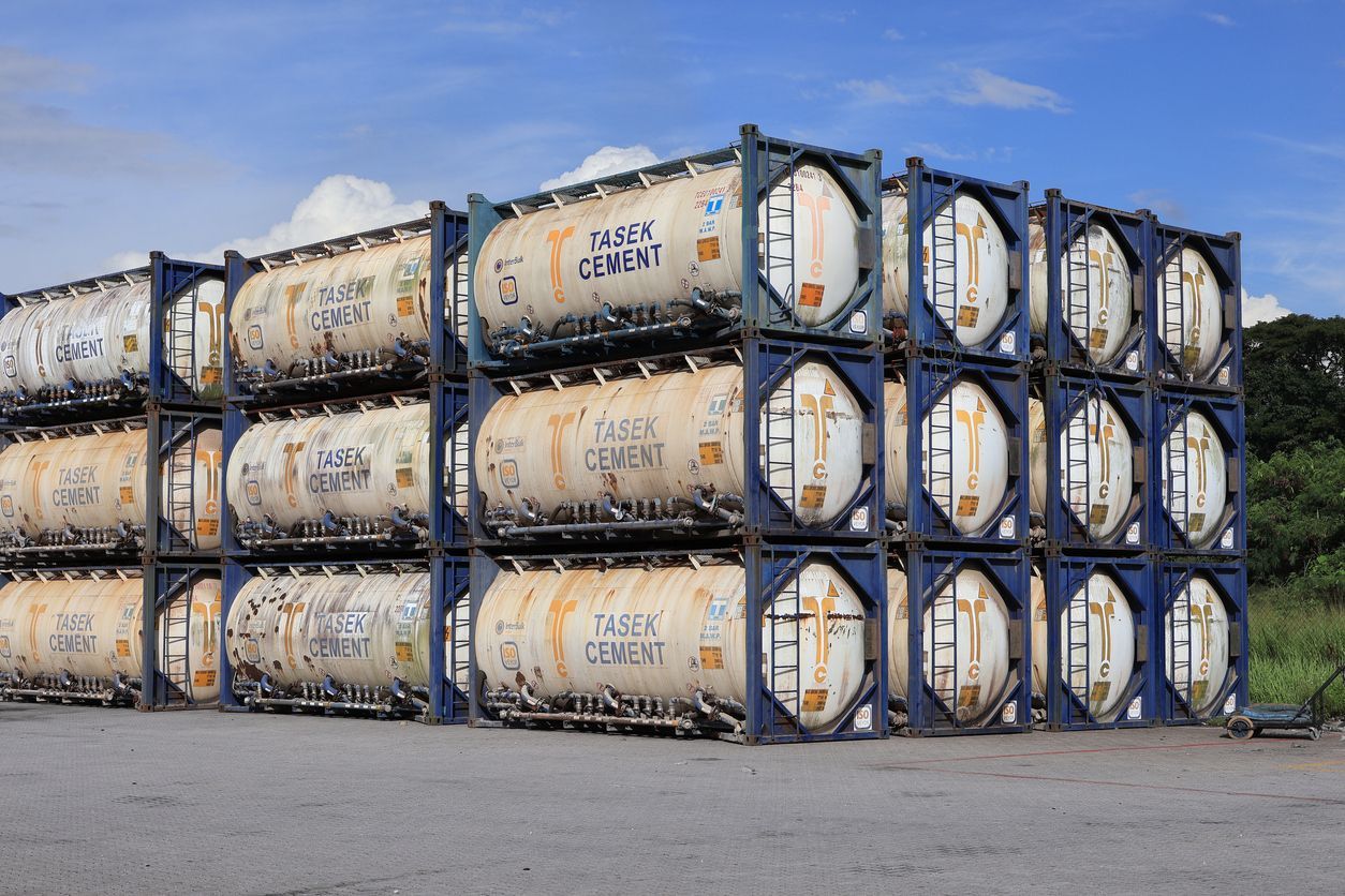 Stacked shipping containers, cylindrical tanks inside blue frames, on a concrete surface, against a blue sky.