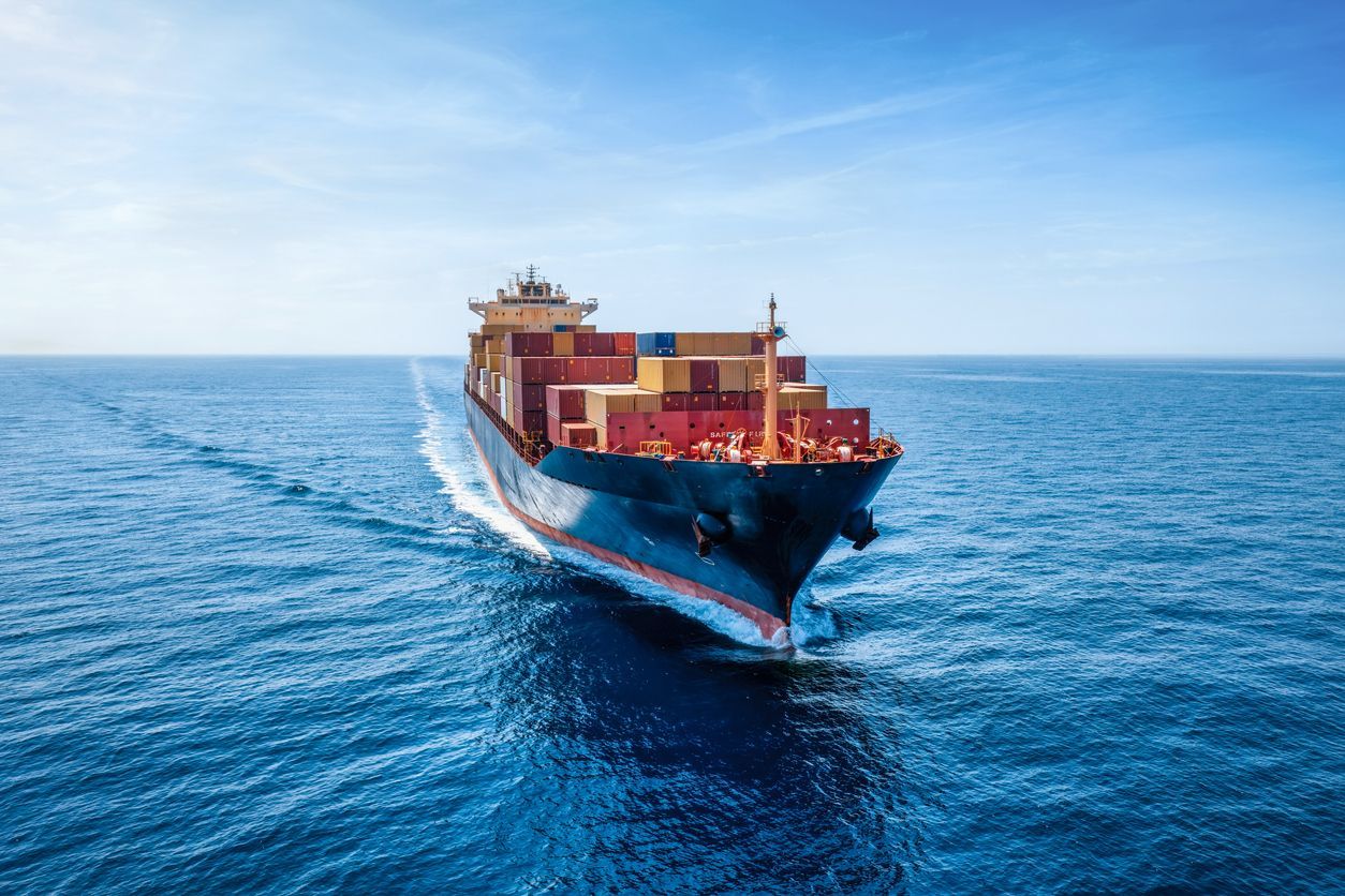 Cargo ship sails across a blue ocean under a clear sky, leaving a wake.