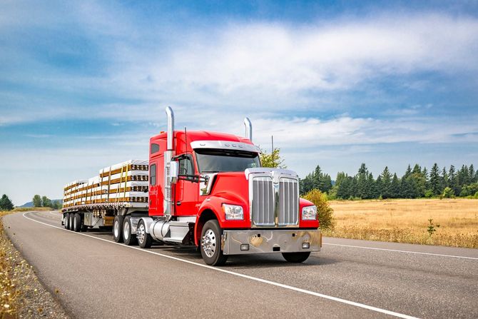 Red semi-truck hauling cargo on a highway under a blue sky with clouds.