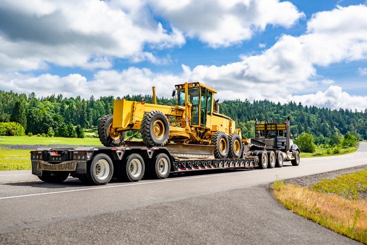 Yellow grader on a flatbed trailer being transported on a road.