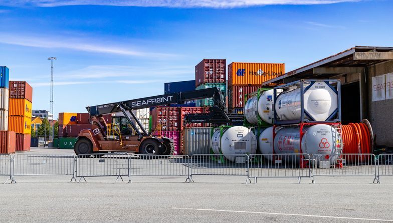 Forklift at a shipping port, moving containers and tanks. Red, blue, orange containers, blue sky.
