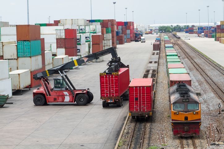 Forklift loading red shipping containers onto a train in a rail yard.