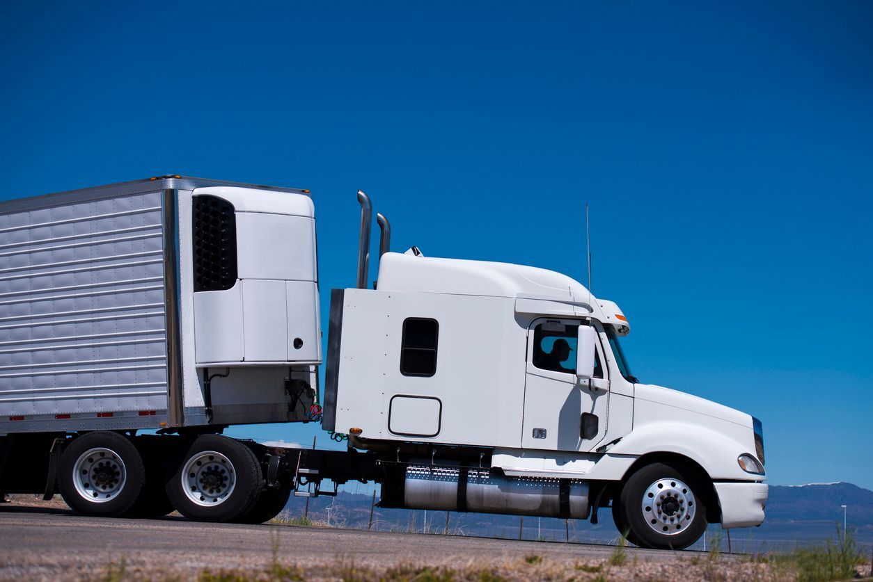 White semi-truck with a refrigerated trailer driving on a road against a clear blue sky.