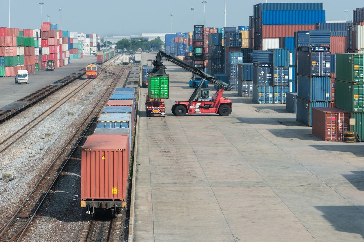 Forklift loading a green shipping container onto a train at a port, surrounded by stacks of colorful containers.