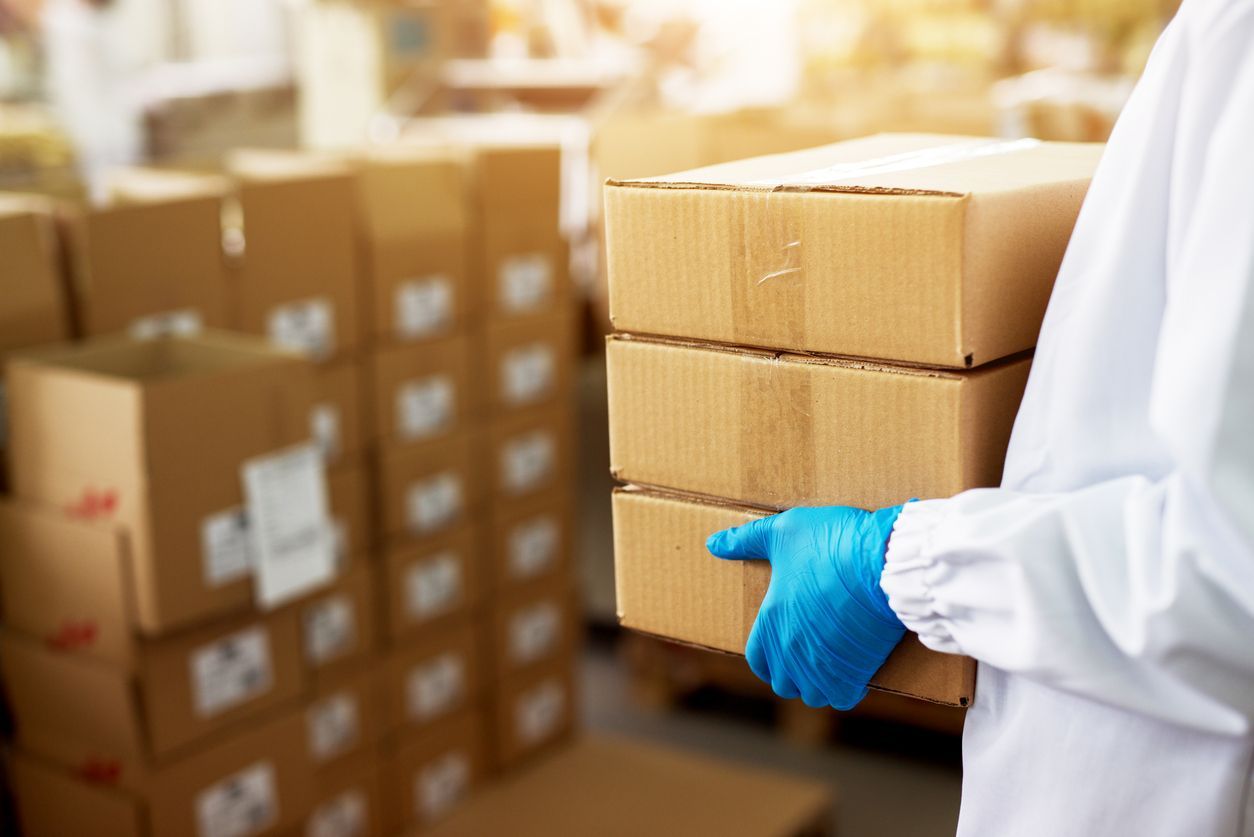 Person wearing gloves and a lab coat carrying three cardboard boxes in a warehouse.