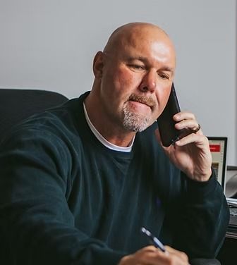 Man on the phone, holding a pen. He has a shaved head, goatee, and is wearing a dark sweater in an office setting.
