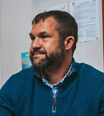 Man in blue sweater, looking to the side, smiling. A full beard and short dark hair.