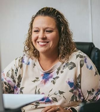 Woman smiling, wearing floral top, seated at a desk.