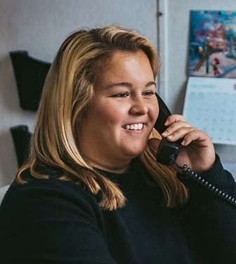 Woman smiles while holding a telephone, sitting in an office, wearing a black shirt.