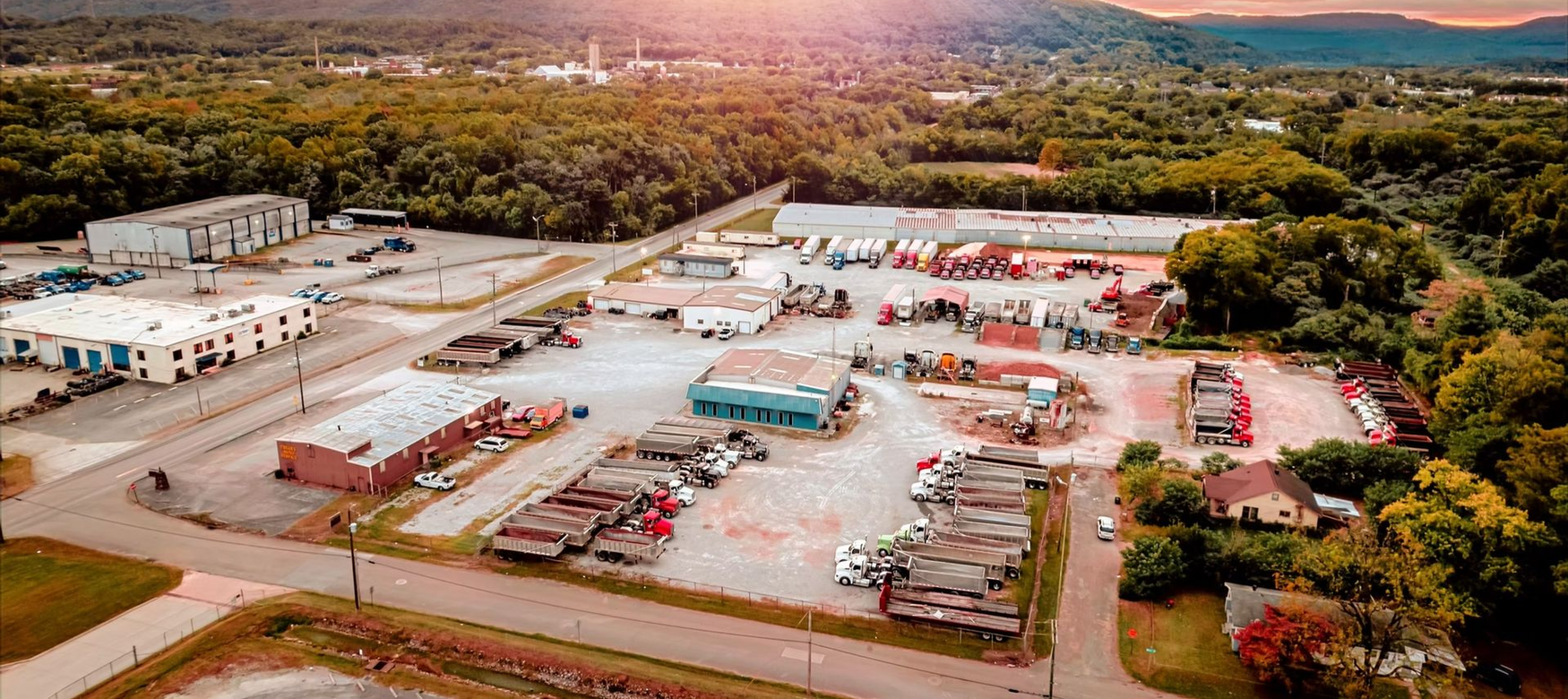 Aerial view of an industrial area with buildings, vehicles, and surrounding trees. Mountain in the background.