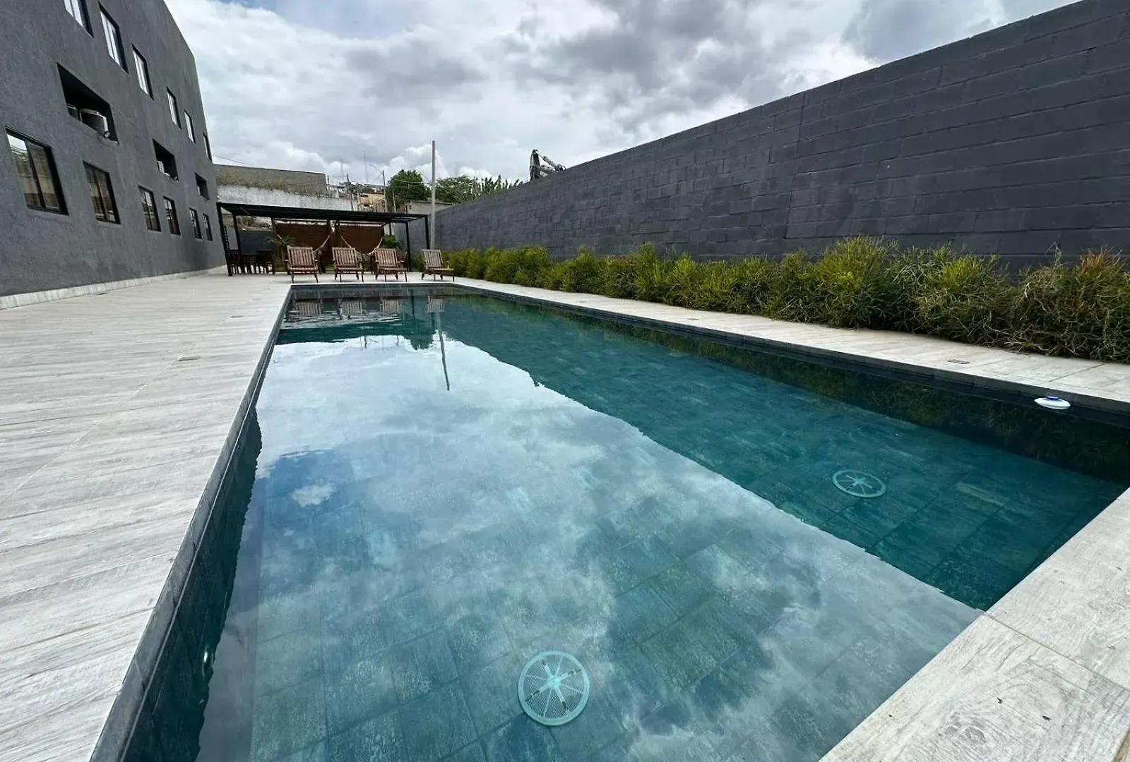 Piscina retangular com água turquesa, rodeada por pedra cinzenta. Ao fundo, uma parede preta e folhagem.
