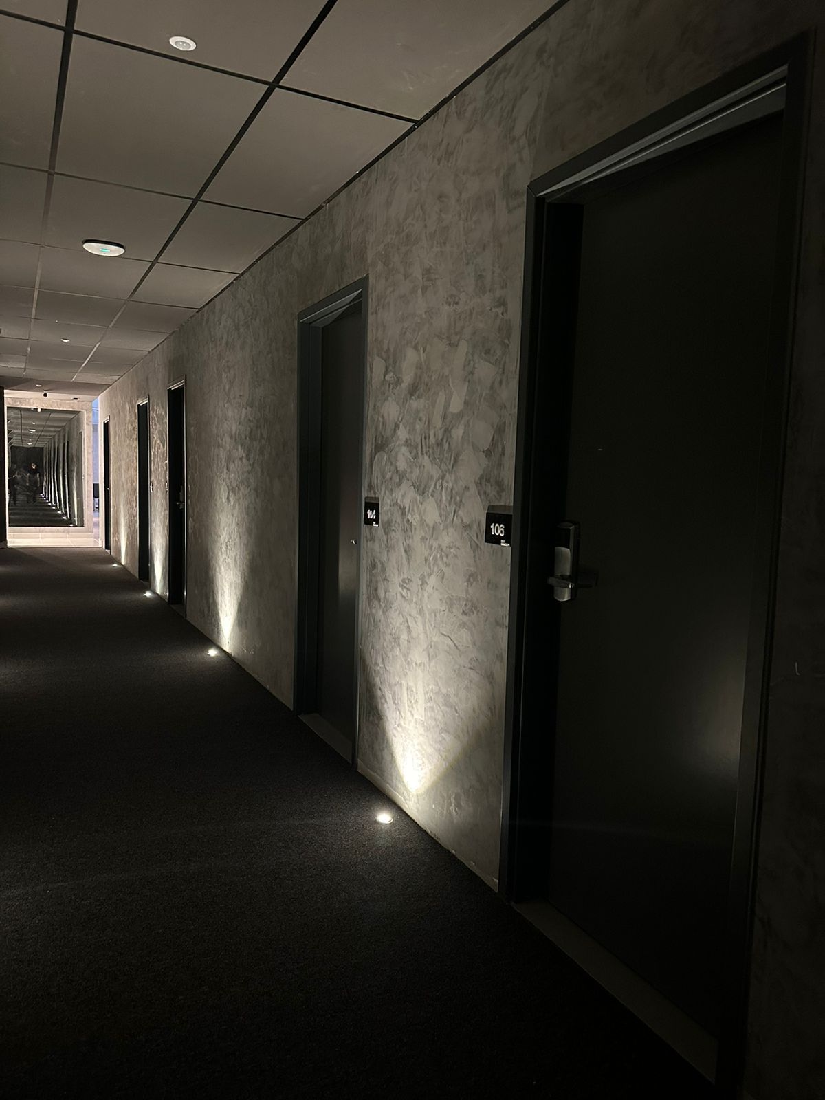 Corredor escuro de hotel com portas e spots de luz em parede texturizada.