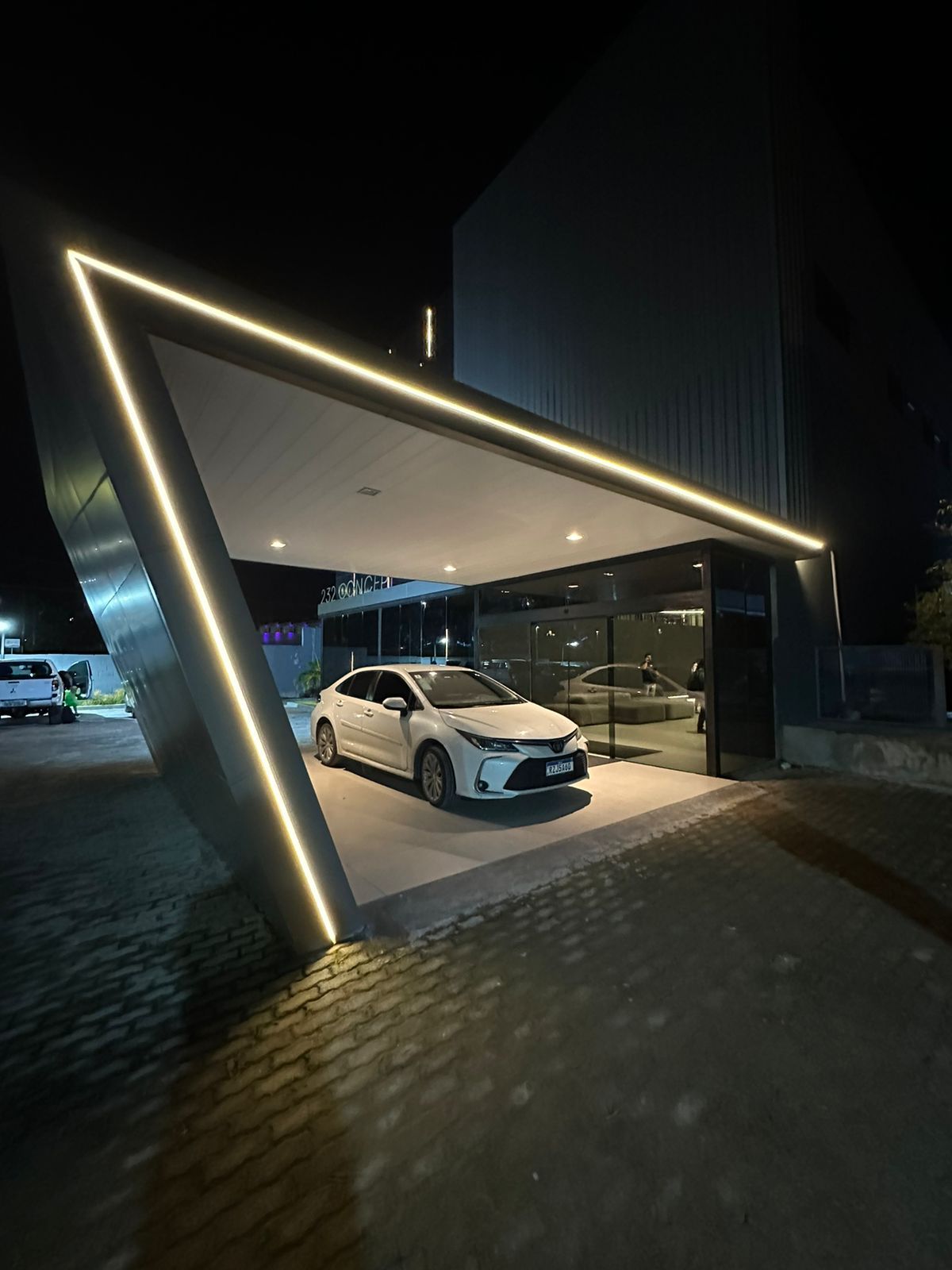 Carro branco estacionado sob uma cobertura triangular moderna com luzes de néon à noite.