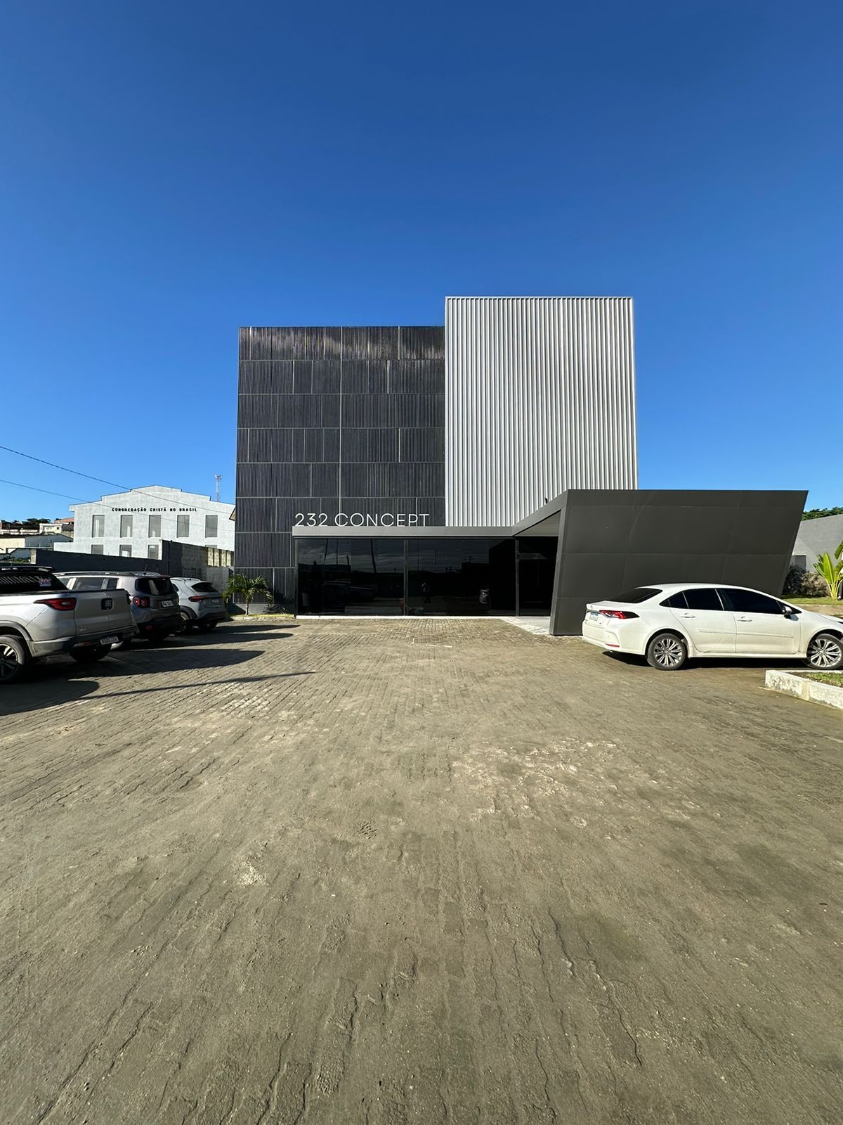Prédio moderno com fachada texturizada em tons de cinza e branco sob um céu azul brilhante. Carros estacionados em um terreno de cascalho.
