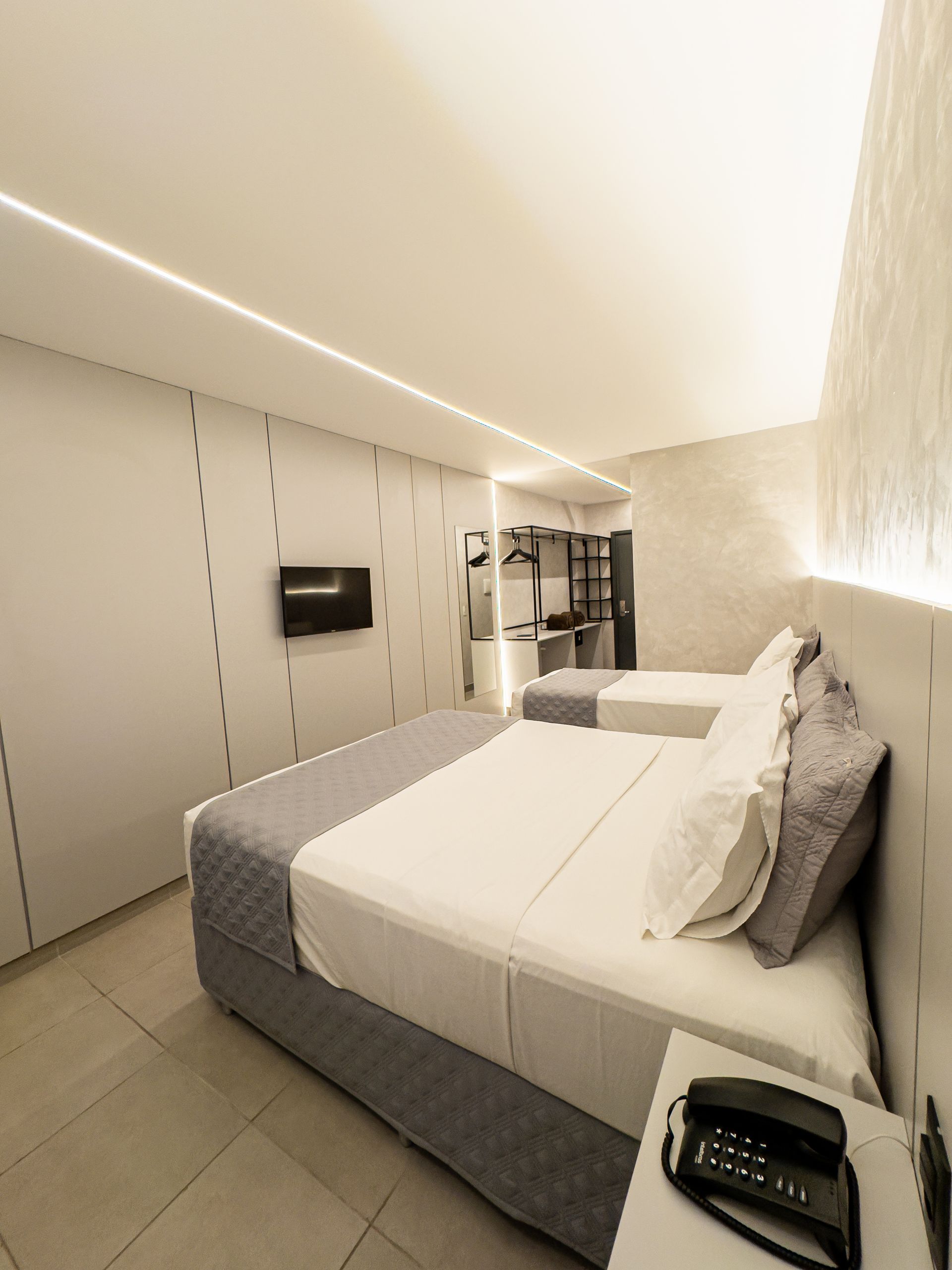 Quarto de hotel com duas camas, paredes brancas e uma TV fixada na parede.