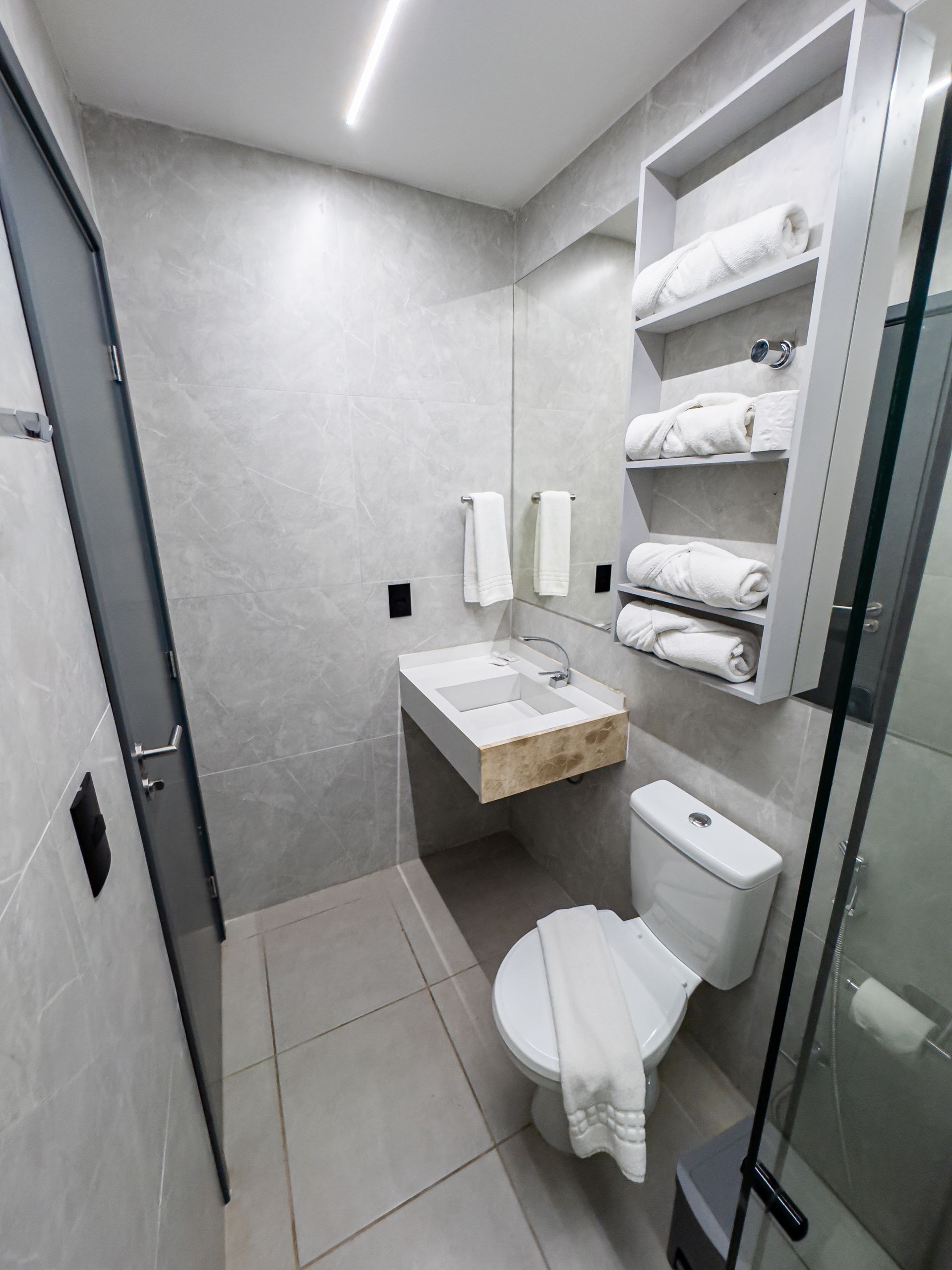 Banheiro pequeno e moderno com paredes revestidas de azulejos cinza, vaso sanitário, pia e porta-toalhas com toalhas brancas.