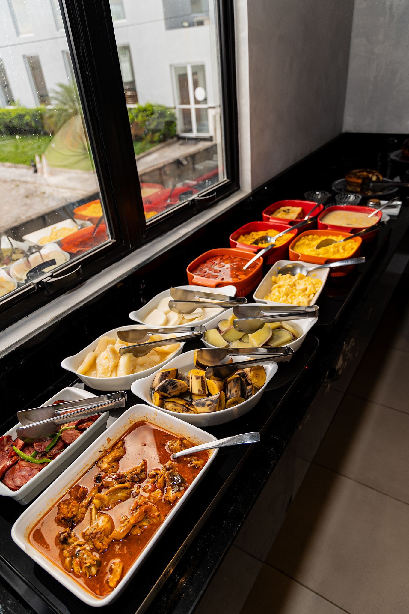 Buffet com diversos pratos servidos em travessas brancas, perto de uma janela.