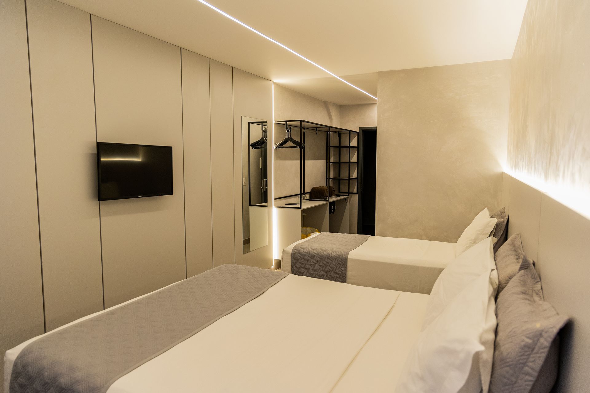 Quarto de hotel moderno com duas camas, TV e armário aberto. Paredes brancas, roupa de cama cinza e iluminação fluorescente.