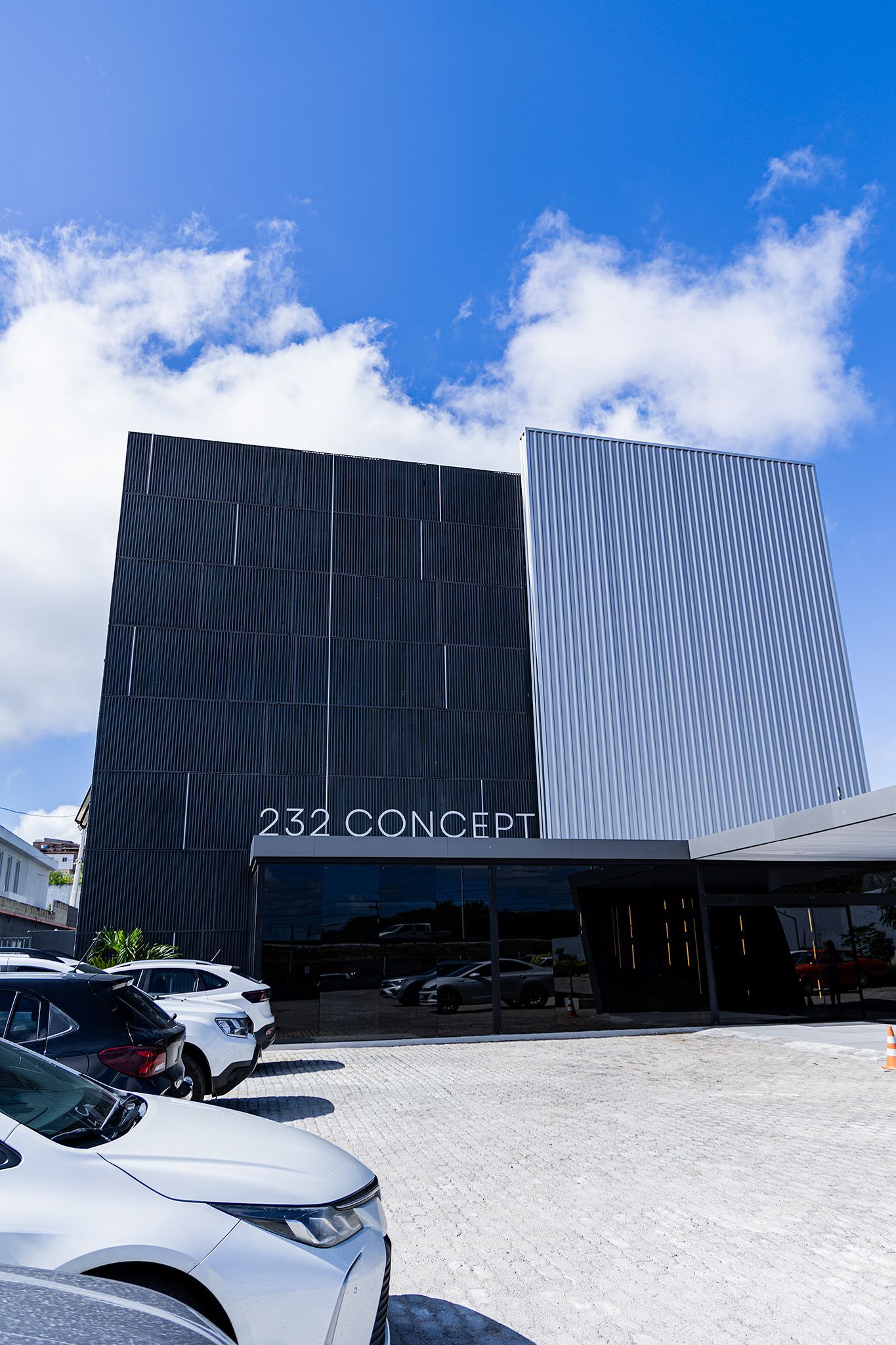 Prédio moderno com fachada preta e prateada, carros estacionados e céu azul.