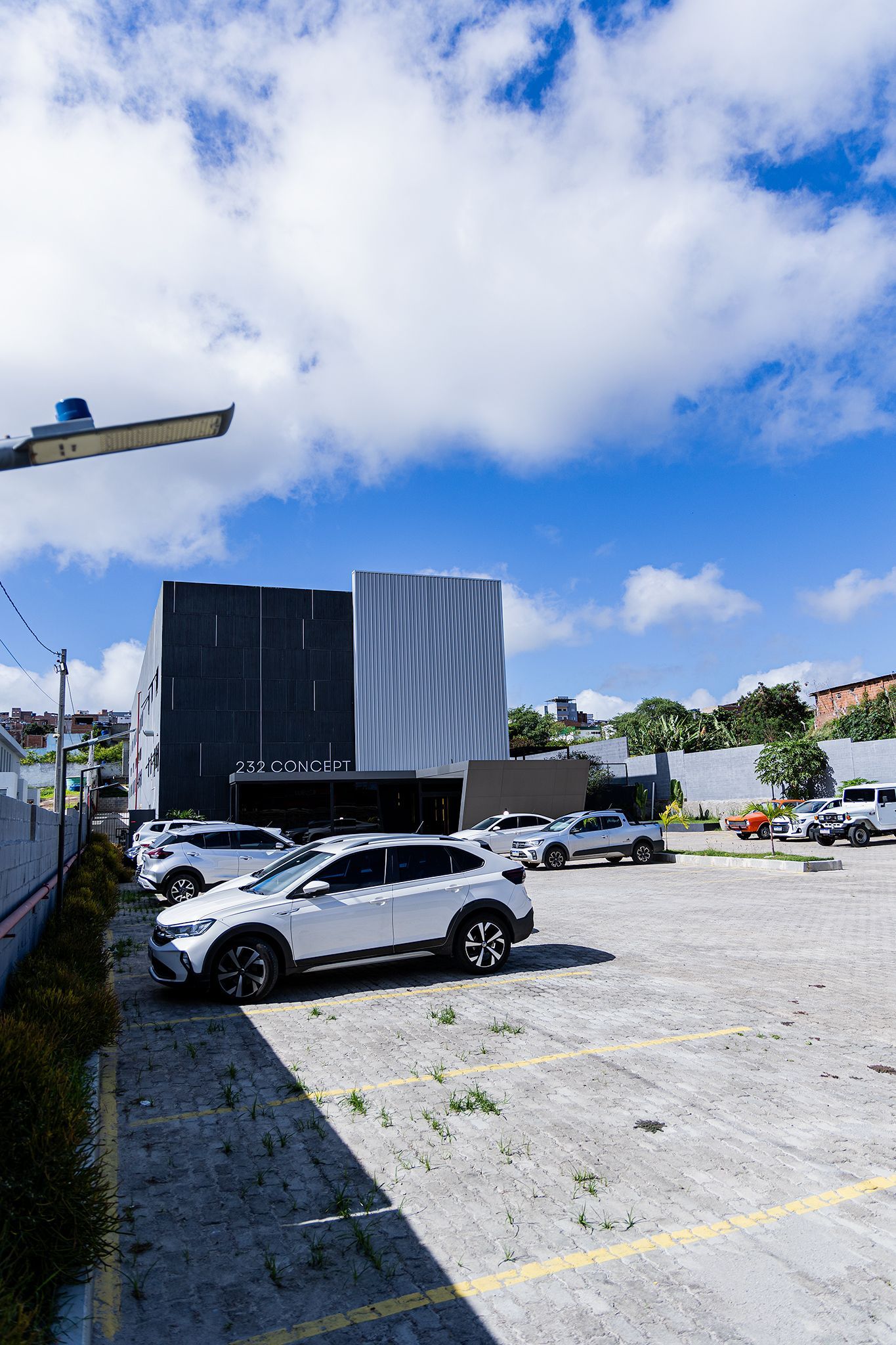 Carros estacionados em um terreno em frente a um prédio moderno, sob um céu azul brilhante com nuvens brancas e fofas.