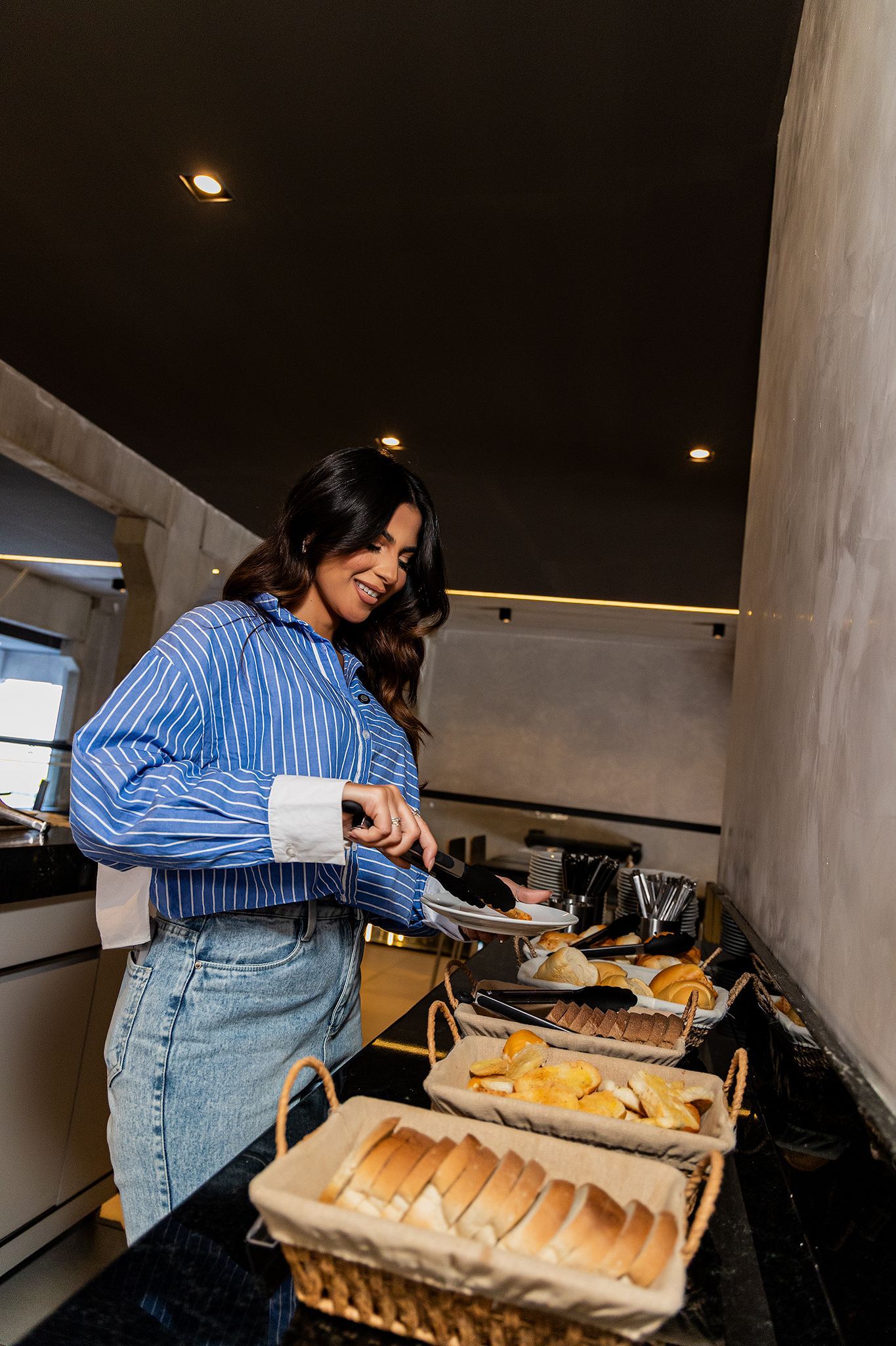 Mulher de camisa azul cortando comida em um bufê. Pães, batatas fritas e outros pratos estão visíveis no balcão.