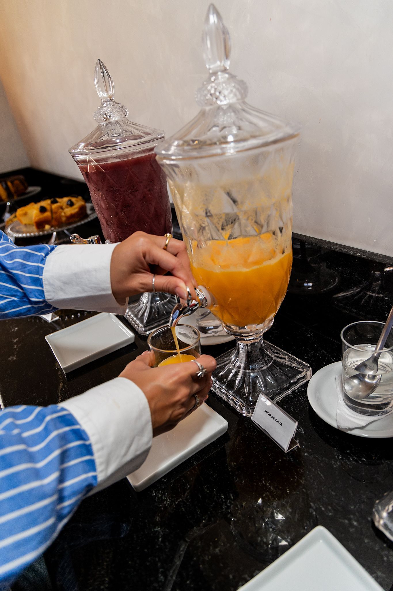 Pessoa servindo suco de laranja de um dispensador. Dois dispensadores de suco sobre uma superfície preta.