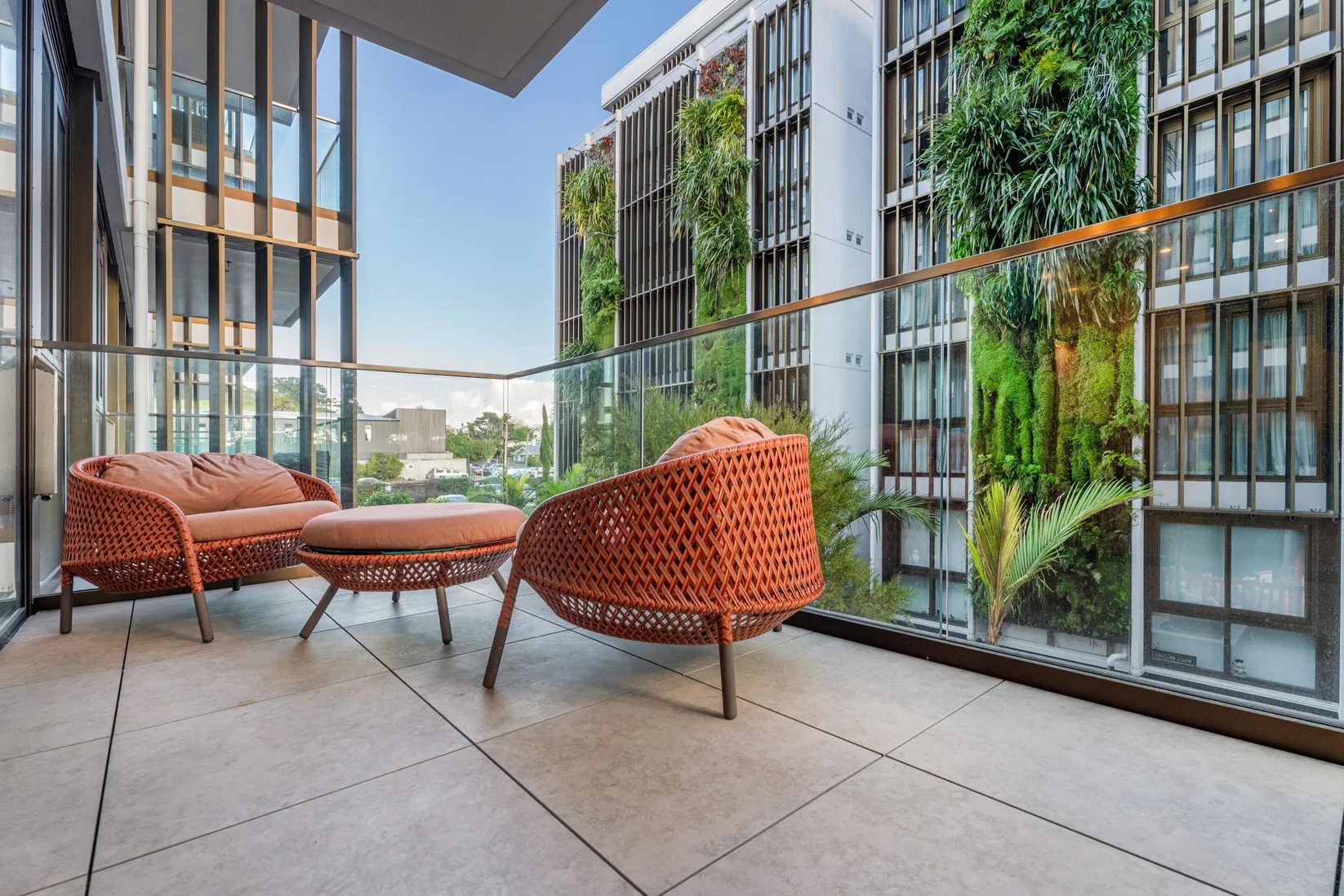 Balcony with orange woven furniture, glass railing, and building with vertical garden.