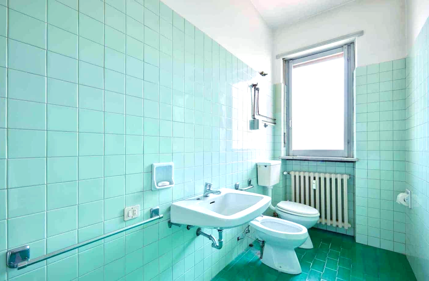 A bathroom with green tiles, a sink, toilet, and window.