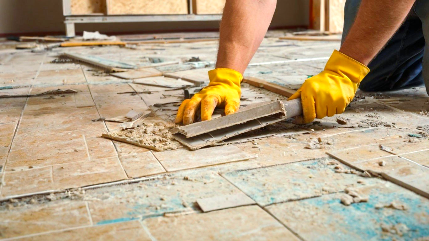 A person wearing yellow gloves is removal tiles from the floor.