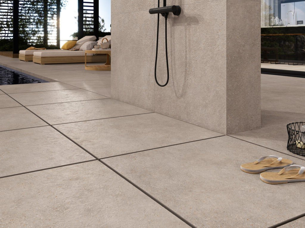 Outdoor shower with tan tiles, black showerhead, and sandals. Pool and lounge chairs in background.