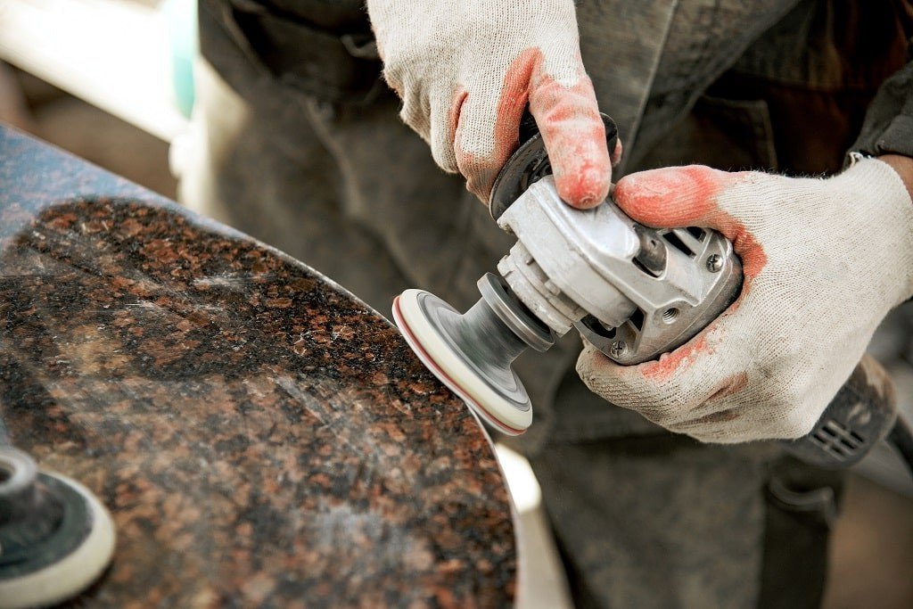 Person wearing gloves using a grinder to polish a brown granite countertop.