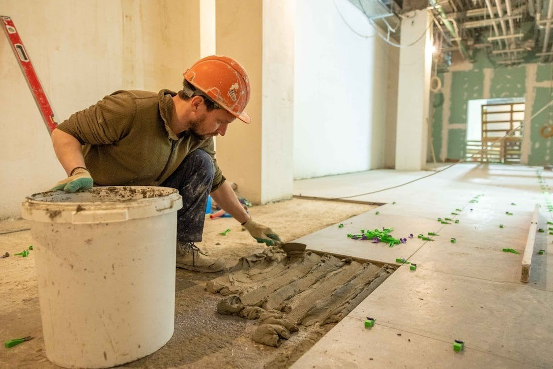 A man wearing a hard hat is laying tiles on a floor.