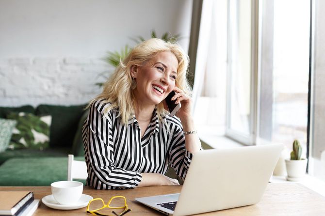 A woman is sitting at a table with a laptop and talking on a cell phone.