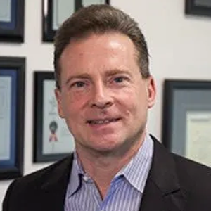 A man in a suit and striped shirt is smiling in front of a wall of framed pictures.