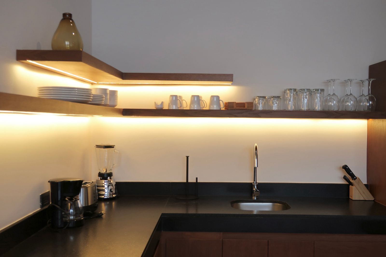 Kitchen with dark countertop, floating wood shelves, and under-shelf lighting. Various dishes and glasses are on the shelves.