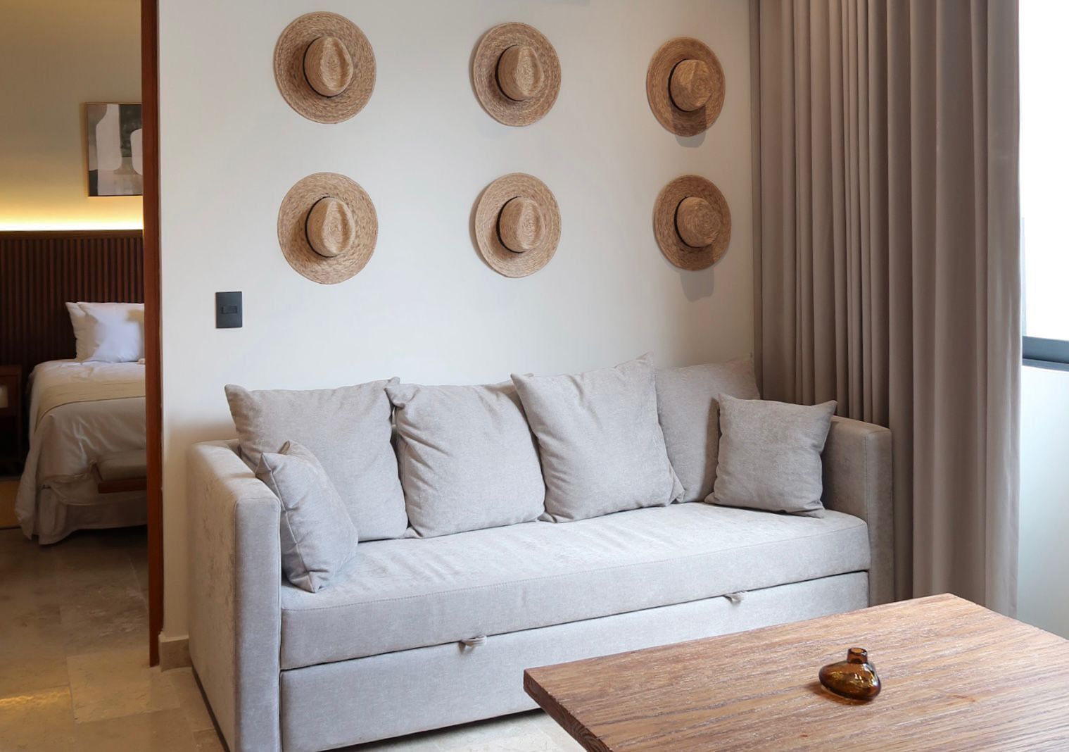 A beige sofa with pillows in a neutral-toned room; straw hats hang above the sofa.