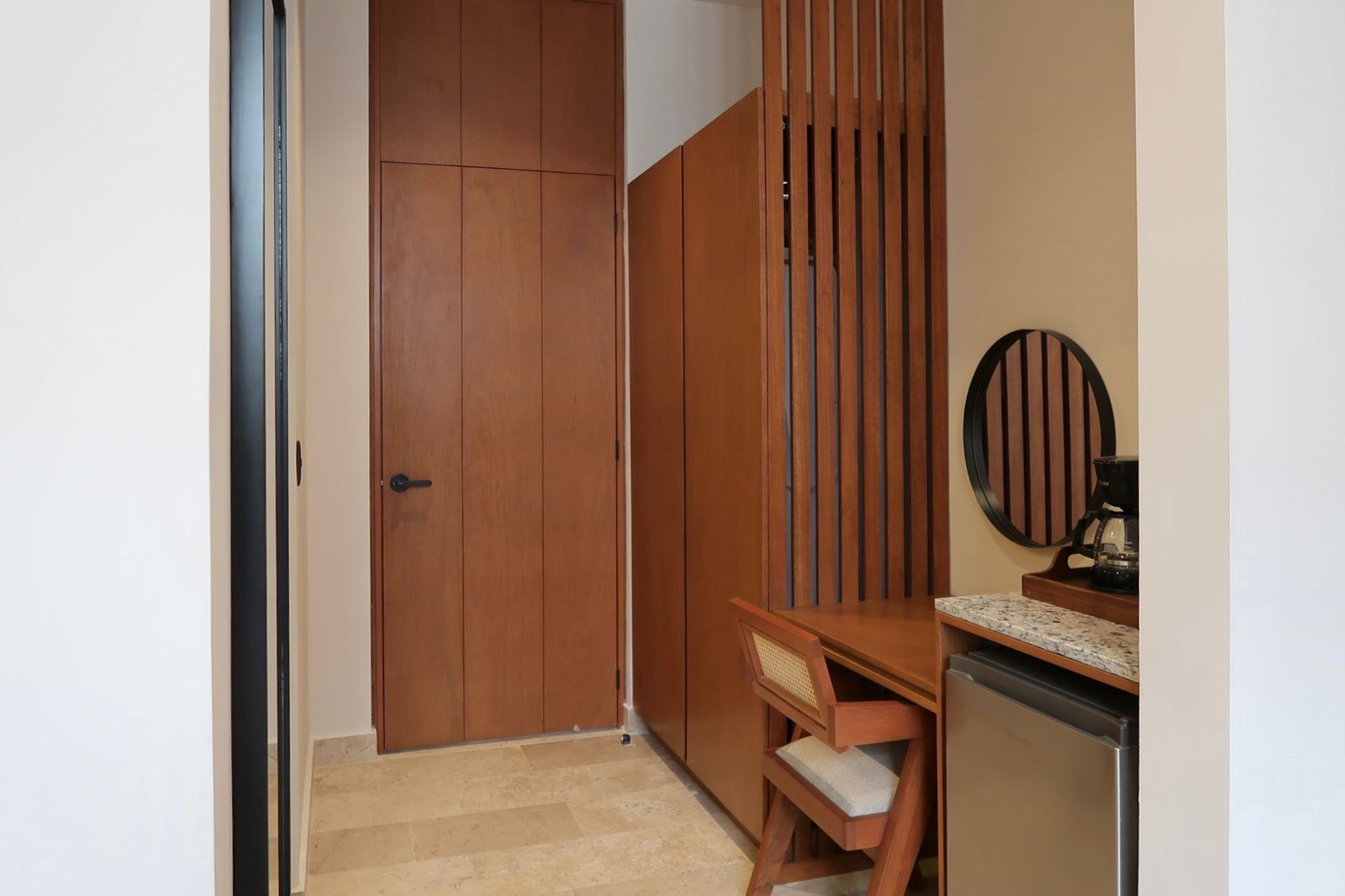 Hallway with brown wooden cabinets, a desk with a mirror and a mini-fridge. Beige walls and stone floor.
