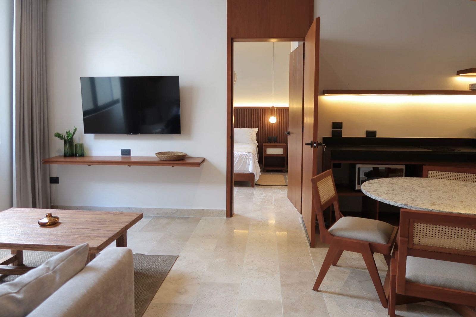 Living room with TV, open door to bedroom, wooden furniture, and light-colored flooring.
