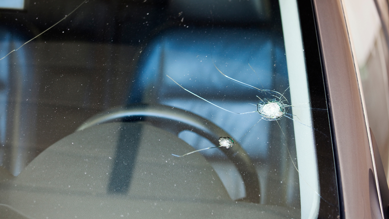 Cracked car windshield with a central impact point and spiderweb-like cracks emanating outward; steering wheel visible.