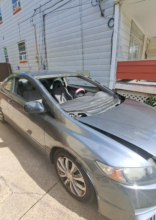 Grey Honda Civic coupe with significant damage to the roof, hood and windshield, parked outside a building.