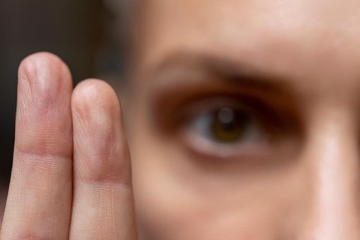 Close-up of a blurred face with a hand raised near the eye, as if peeking through fingers