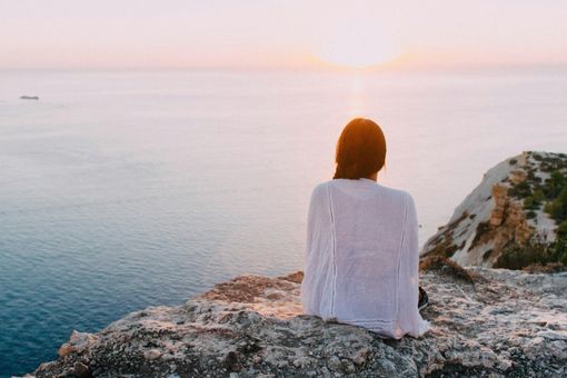 Person sitting on a rocky cliff at sunset, facing a calm ocean.