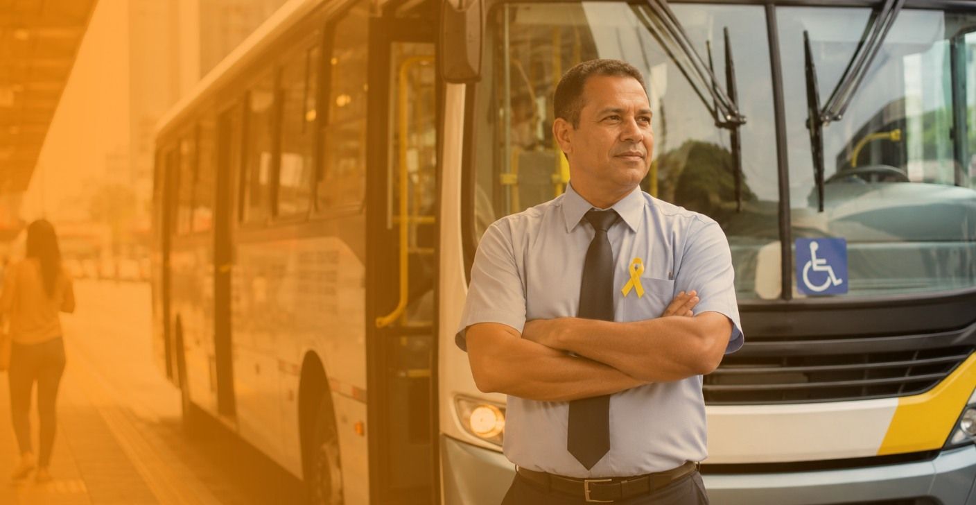 Motorista de ônibus em pé, com os braços cruzados em frente a um ônibus.