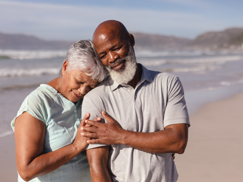Couple embracing on a sunny beach; woman leans on man's shoulder.