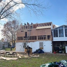 Construction on a two-story building with workers on the roof, framing and boarding the structure, sunny day.
