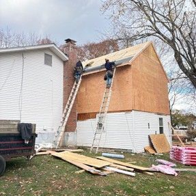 Two roofers on ladders working on a house with exposed plywood. Lumber and materials lay on the ground.